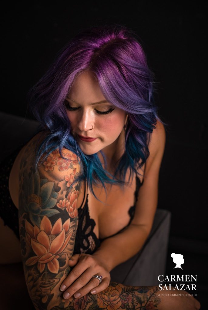 woman with colorful tattoos and purple and blue hair in black lacy lingerie with black background; Boudoir Photography by Carmen Salazar