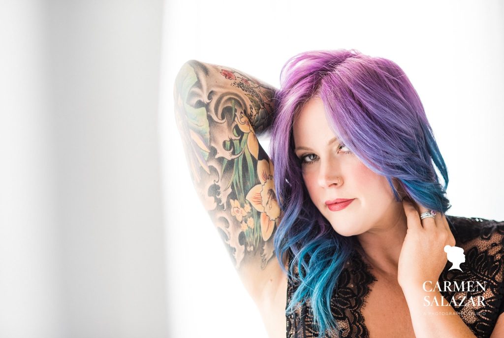 woman with colorful tattoos and purple and blue hair in black lacy lingerie with white background; Boudoir Photography by Carmen Salazar