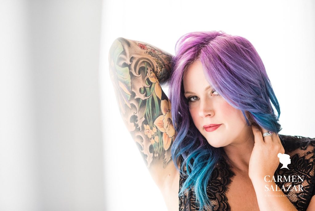 woman with colorful tattoos and purple and blue hair in black lacy lingerie with white background; Boudoir Photography by Carmen Salazar 